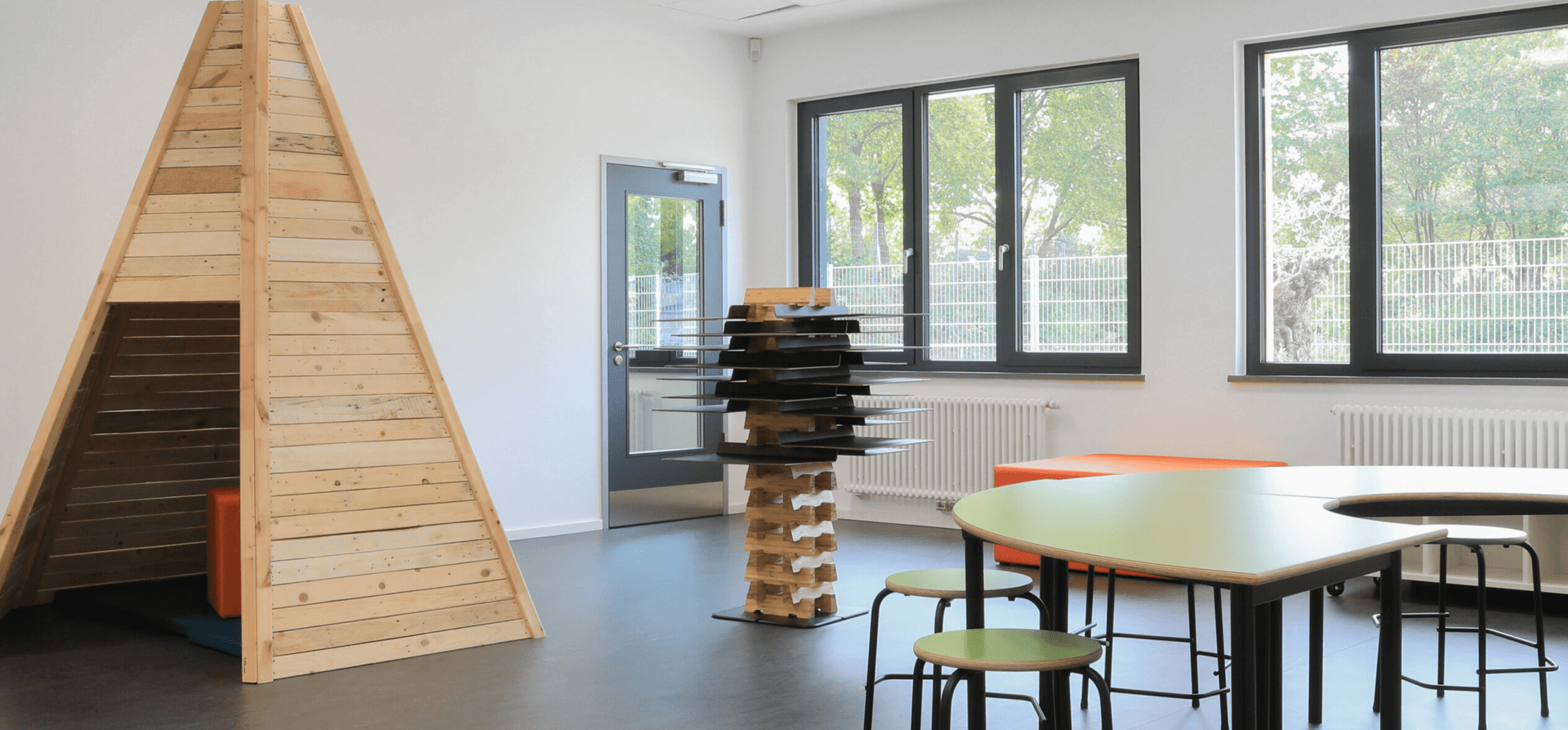 Klassenraum mit großen Fensterflächen, möbliert mit grünen Tischen und einem Tippi aus Holz